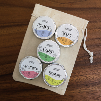Five small round silver foil-wrapped shower steamers labeled Peace, Arise, Ease, Embrace, and Restore from Simply Healthy, arranged on a cream drawstring pouch over a wooden surface.