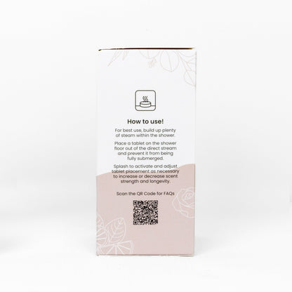 The back of a white product box of Shower Steamer Treat Pack with a light pink lower section, featuring a 'How to use!' icon, instructions, a QR code, and faint floral line art on a white background.